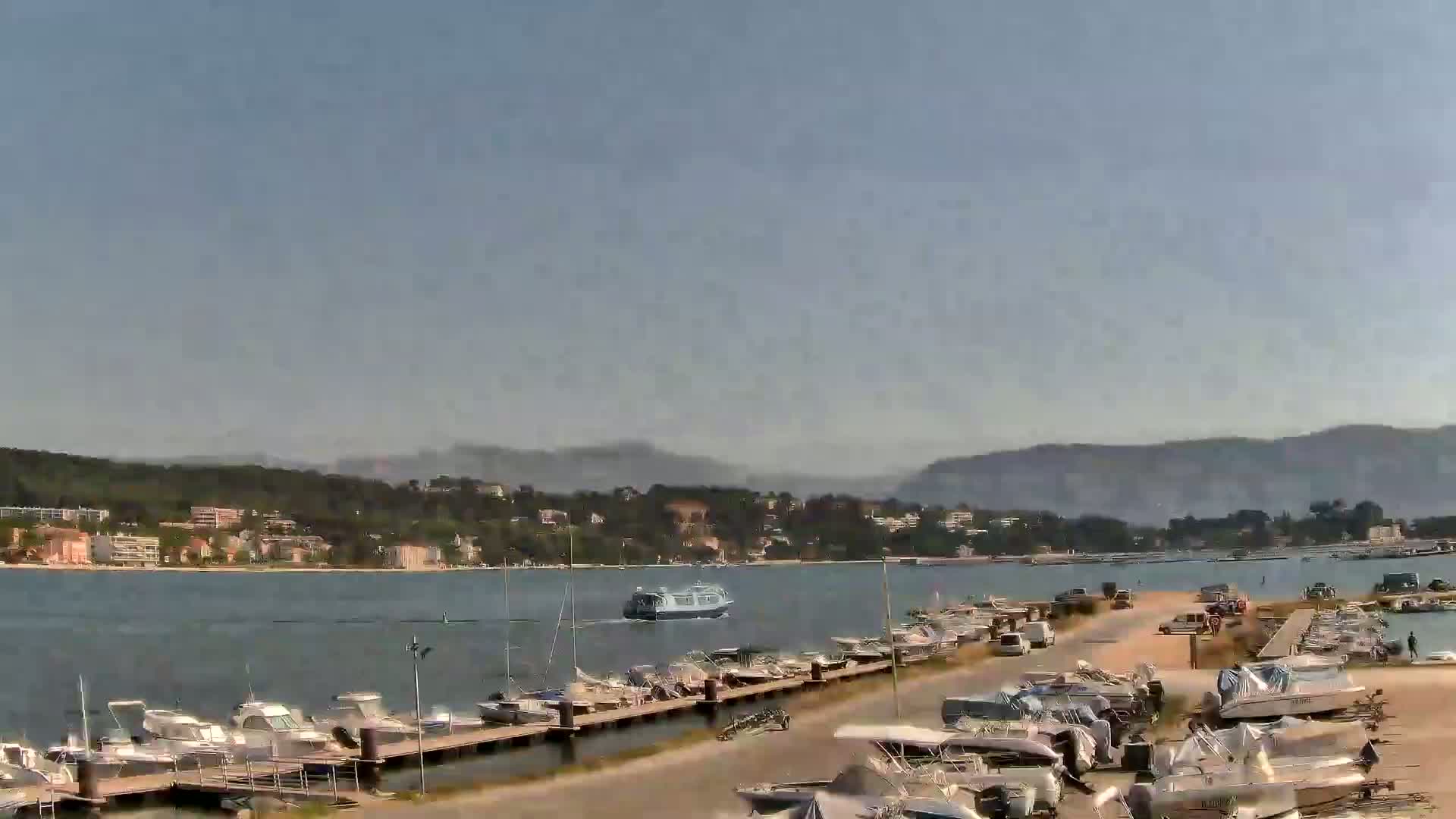 A marina filled with numerous boats is situated near a calm body of water, with a ferry in the distance and hills under a clear sky in the background.