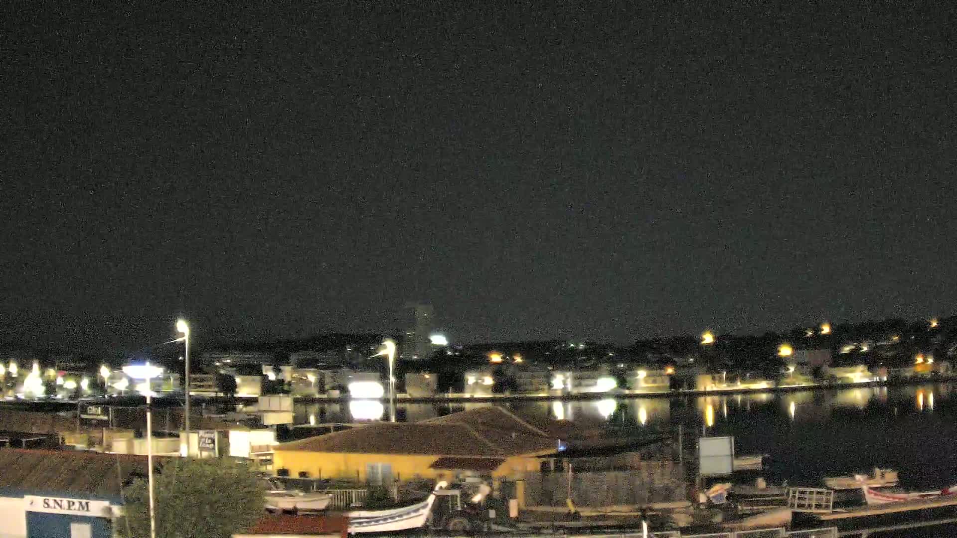 A nighttime view of a calm body of water reflecting the lights of buildings and boats on the shore under a clear, dark sky.