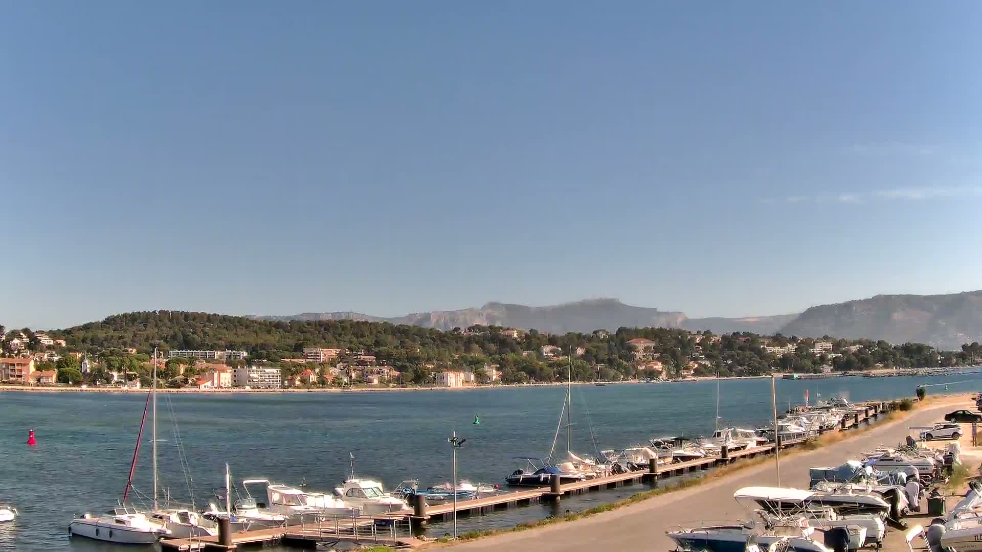A calm, sunny day shows a marina filled with docked boats, a shoreline of buildings and trees, and mountains in the distance.