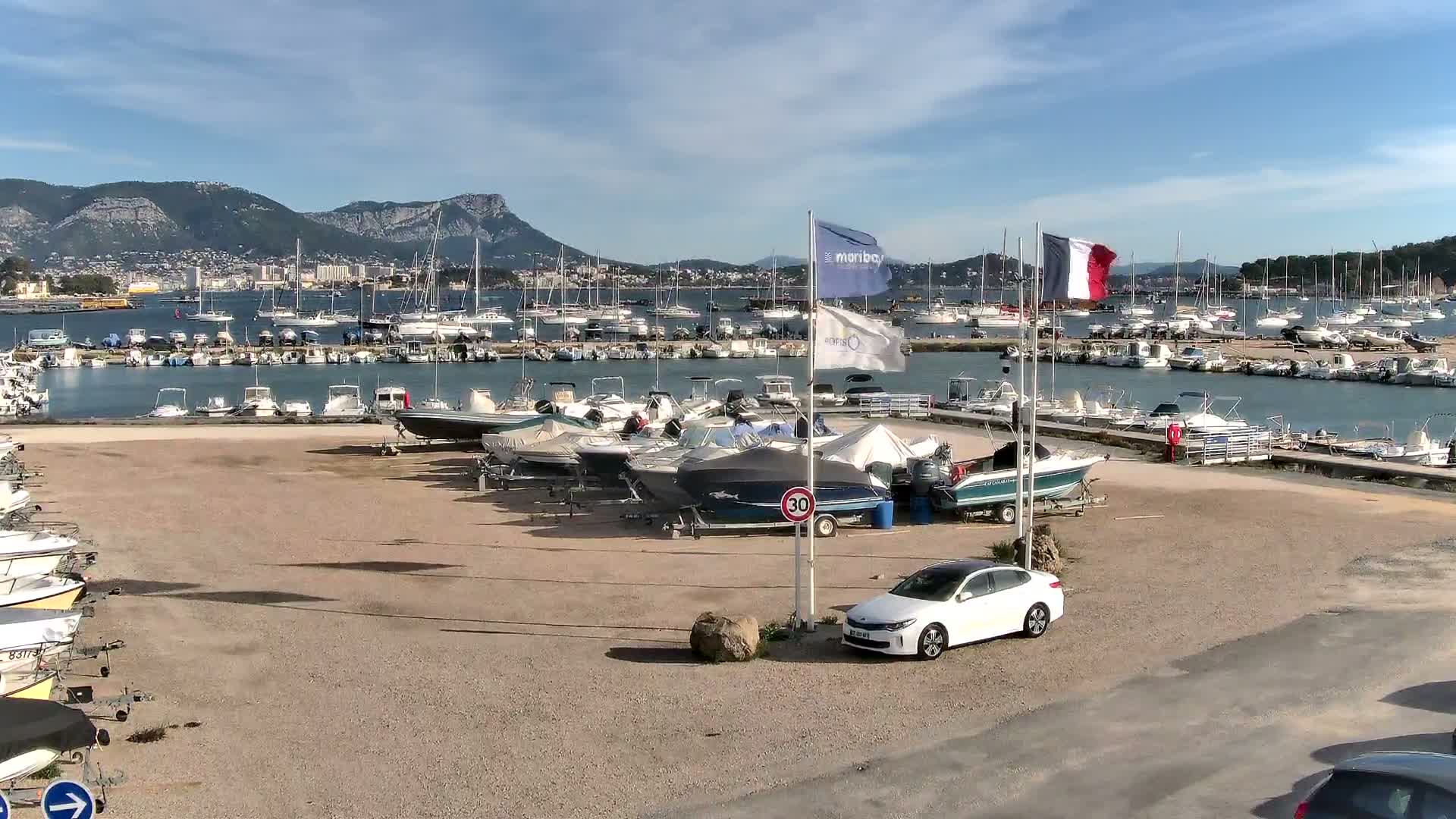 A bright, partly cloudy day illuminates a bustling marina filled with numerous boats afloat and stored ashore, framed by distant mountains and a coastal city, with a white sedan parked prominently in the foreground.
