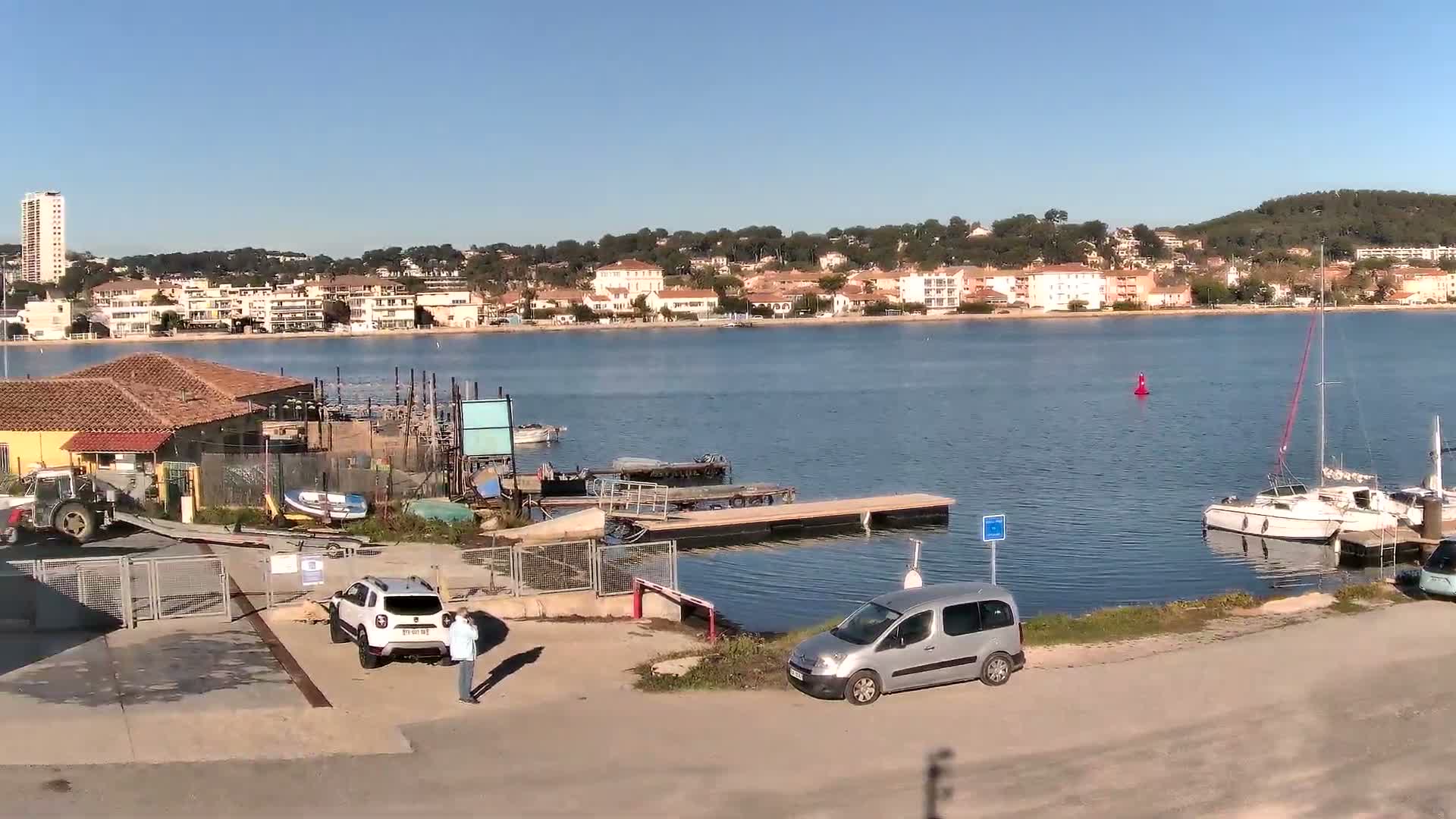 On a clear and sunny day, a tranquil bay with boats and a wooden dock separates a busy foreground featuring a boatyard, parked vehicles, and a person, from a distant coastal town built along green hills.