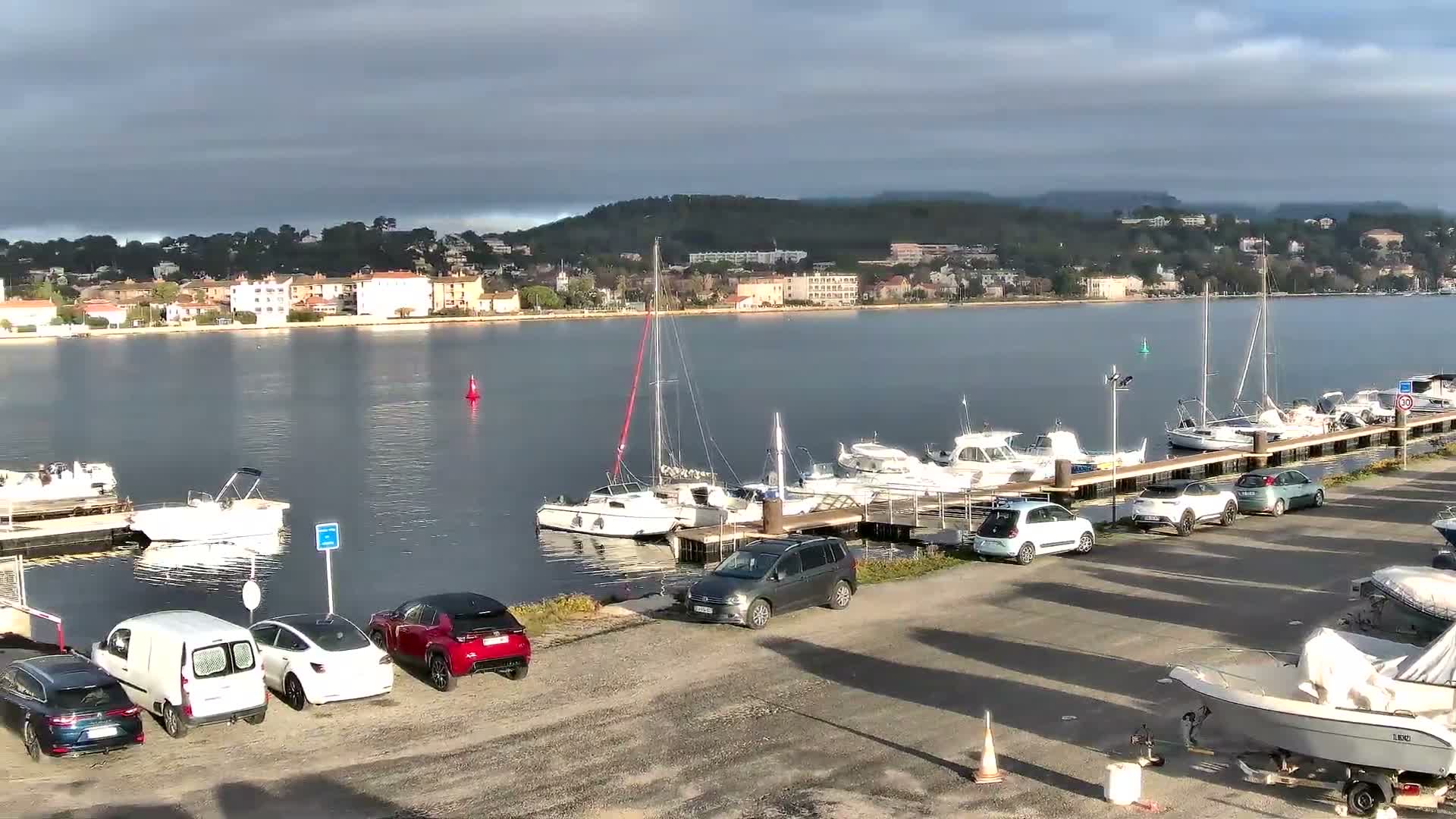 Calm waters of a harbor reflect a partly cloudy sky with hints of sunlight, showing numerous boats docked along piers, several cars parked on a paved lot in the foreground, and a town with verdant hills in the distance.