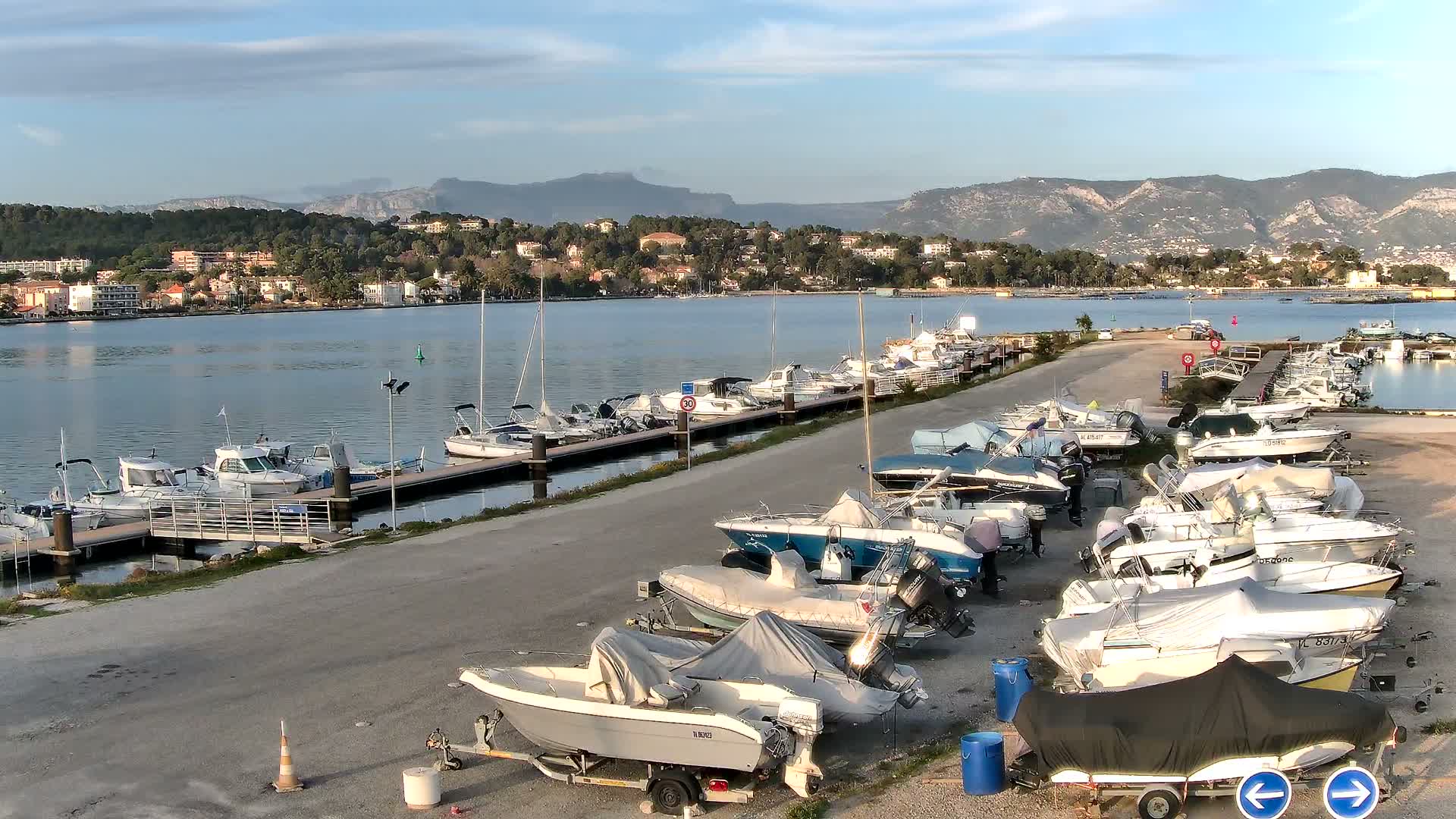 Calm waters of a harbor reflect a partly cloudy sky with hints of sunlight, showing numerous boats docked along piers, several cars parked on a paved lot in the foreground, and a town with verdant hills in the distance.