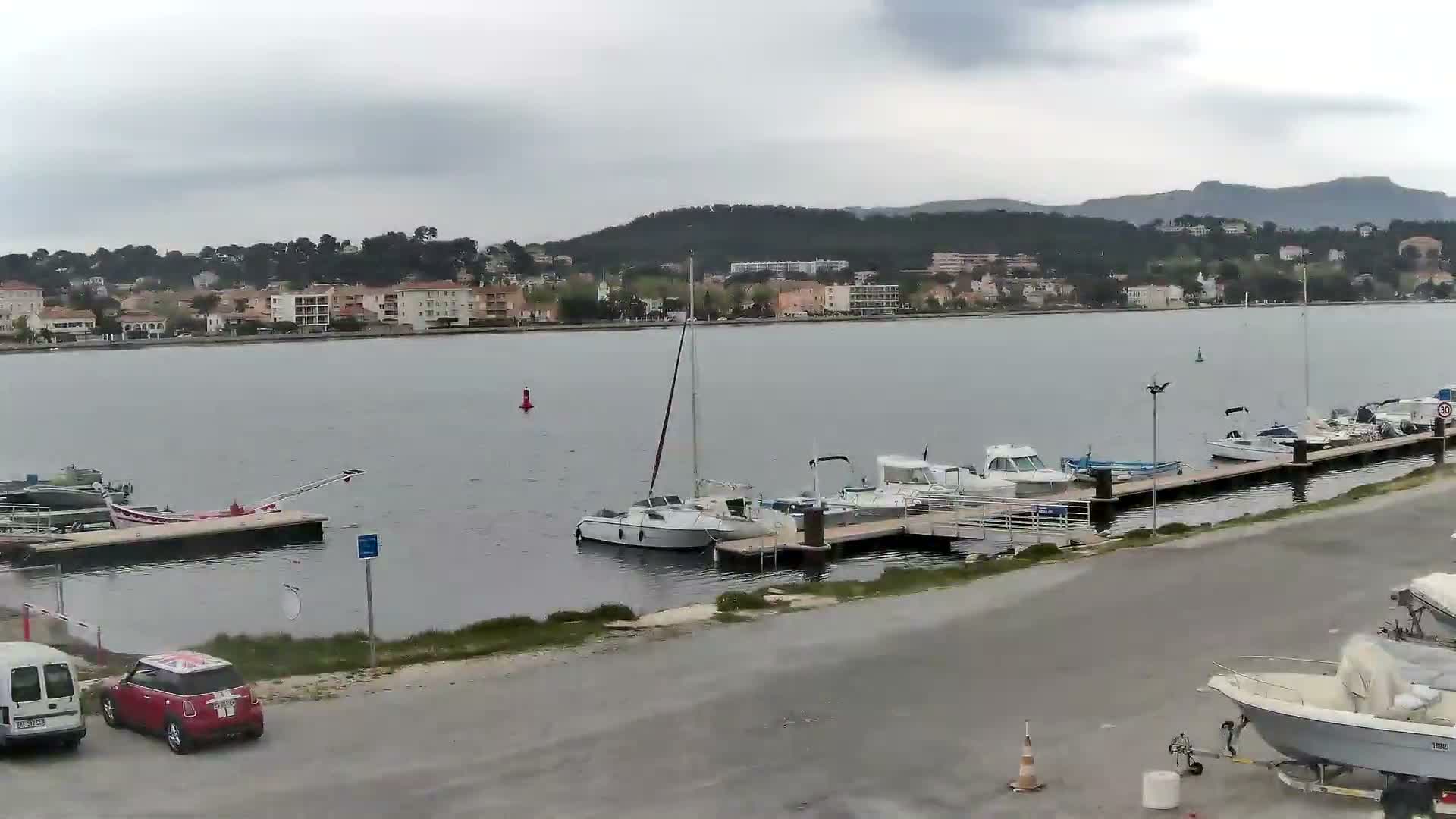 Calm waters of a harbor reflect a partly cloudy sky with hints of sunlight, showing numerous boats docked along piers, several cars parked on a paved lot in the foreground, and a town with verdant hills in the distance.