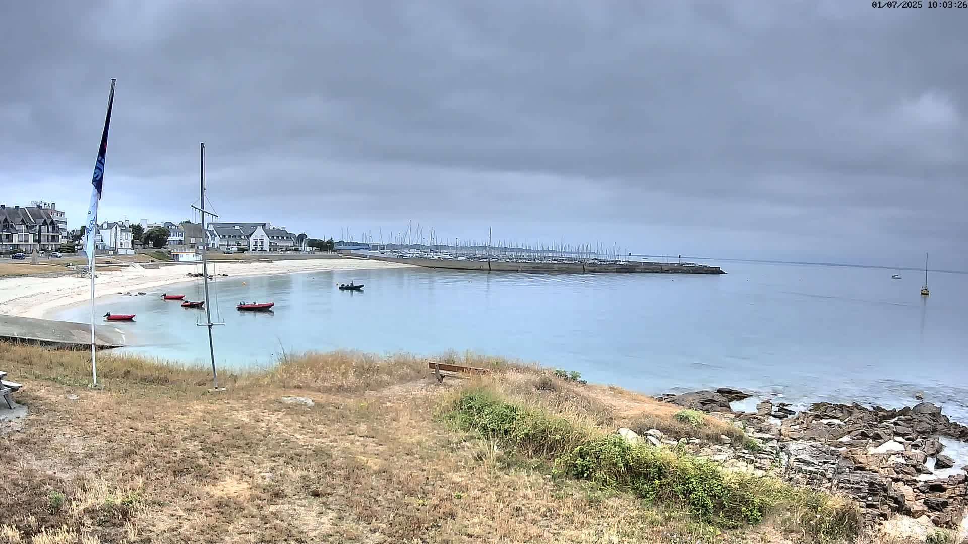 A sandy beach with several small boats in calm, light-blue water under an overcast sky,  backed by buildings and a marina full of sailboats.