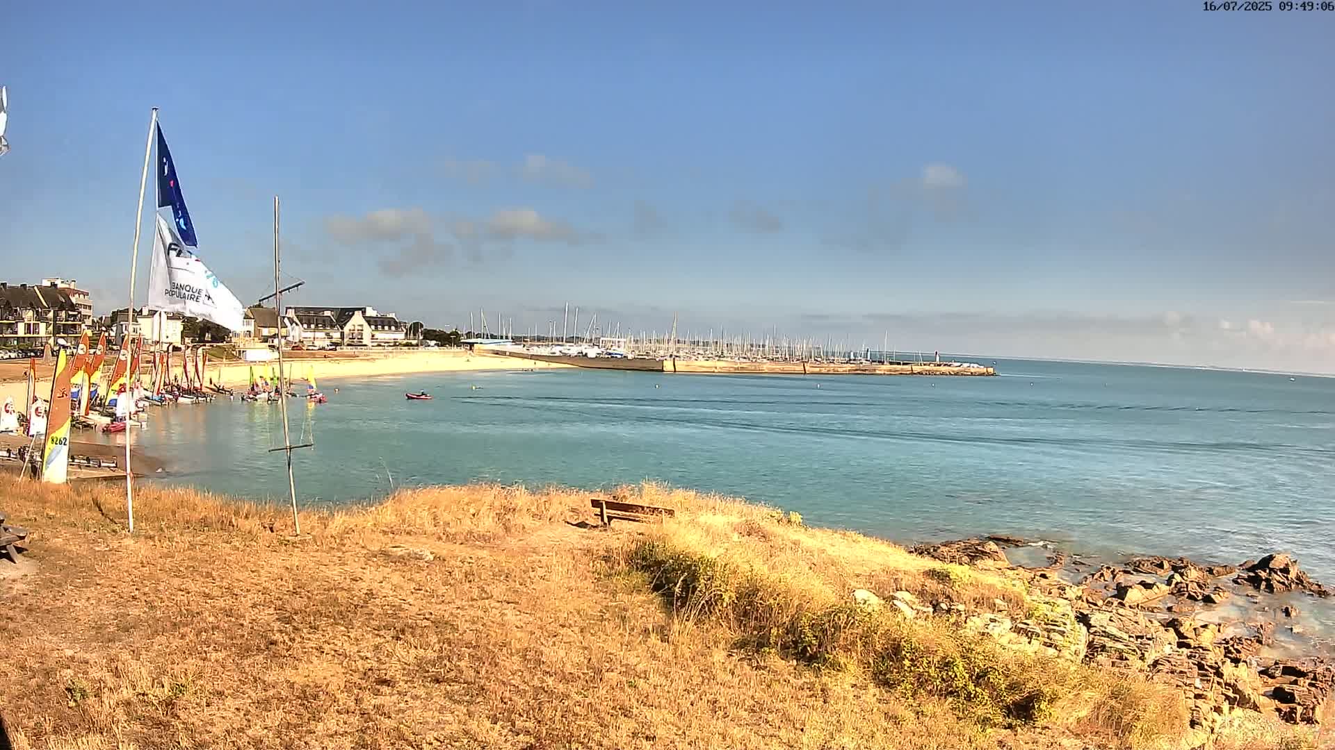 A calm, sunny day shows a harbor filled with sailboats, a sandy beach, and a rocky shoreline with dry, tan grass.