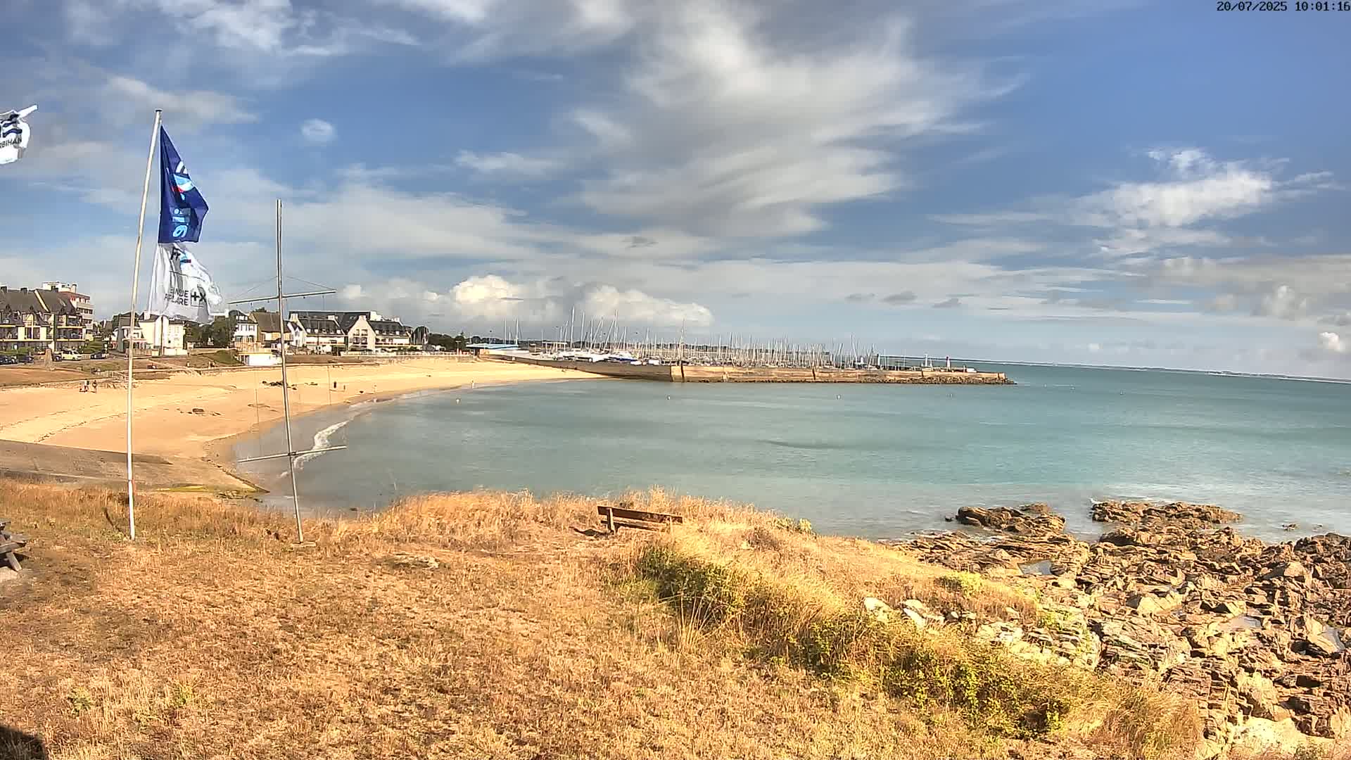 A sandy beach curves into a calm, light-blue ocean harboring numerous sailboats under a partly cloudy sky.