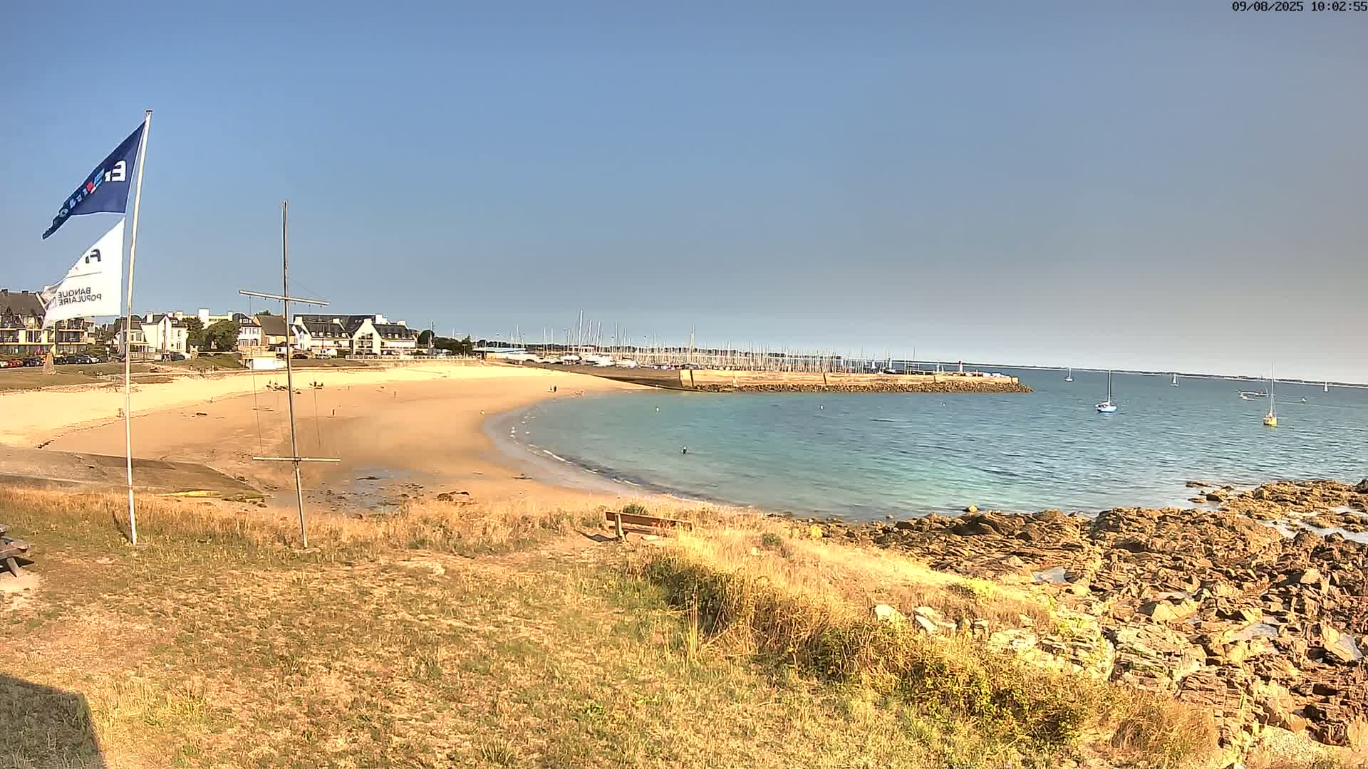A sandy beach curves into a calm, teal ocean, with a marina and buildings visible in the background under a clear blue sky.