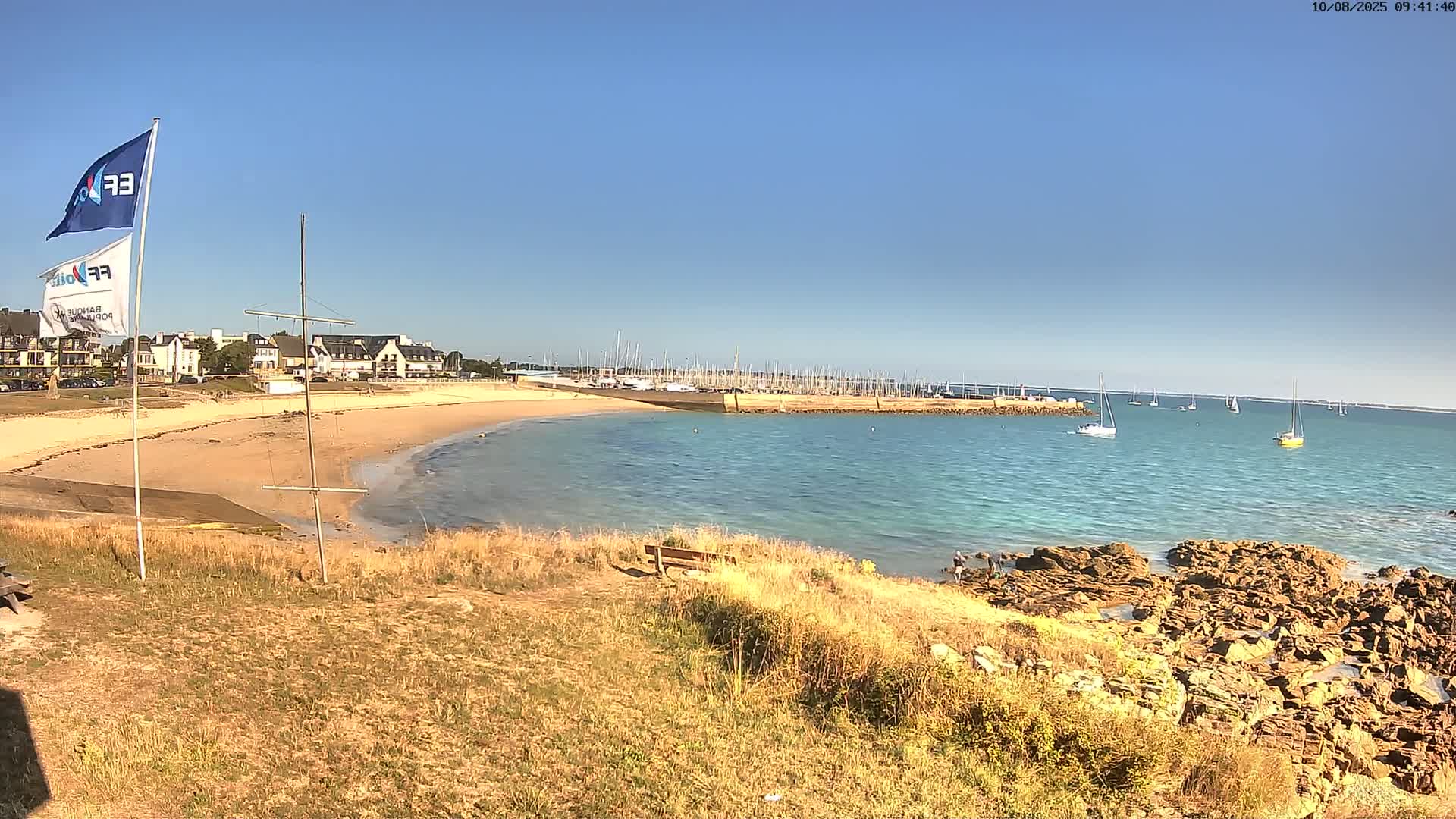 A sandy beach curves around a calm, turquoise bay filled with sailboats under a clear blue sky.