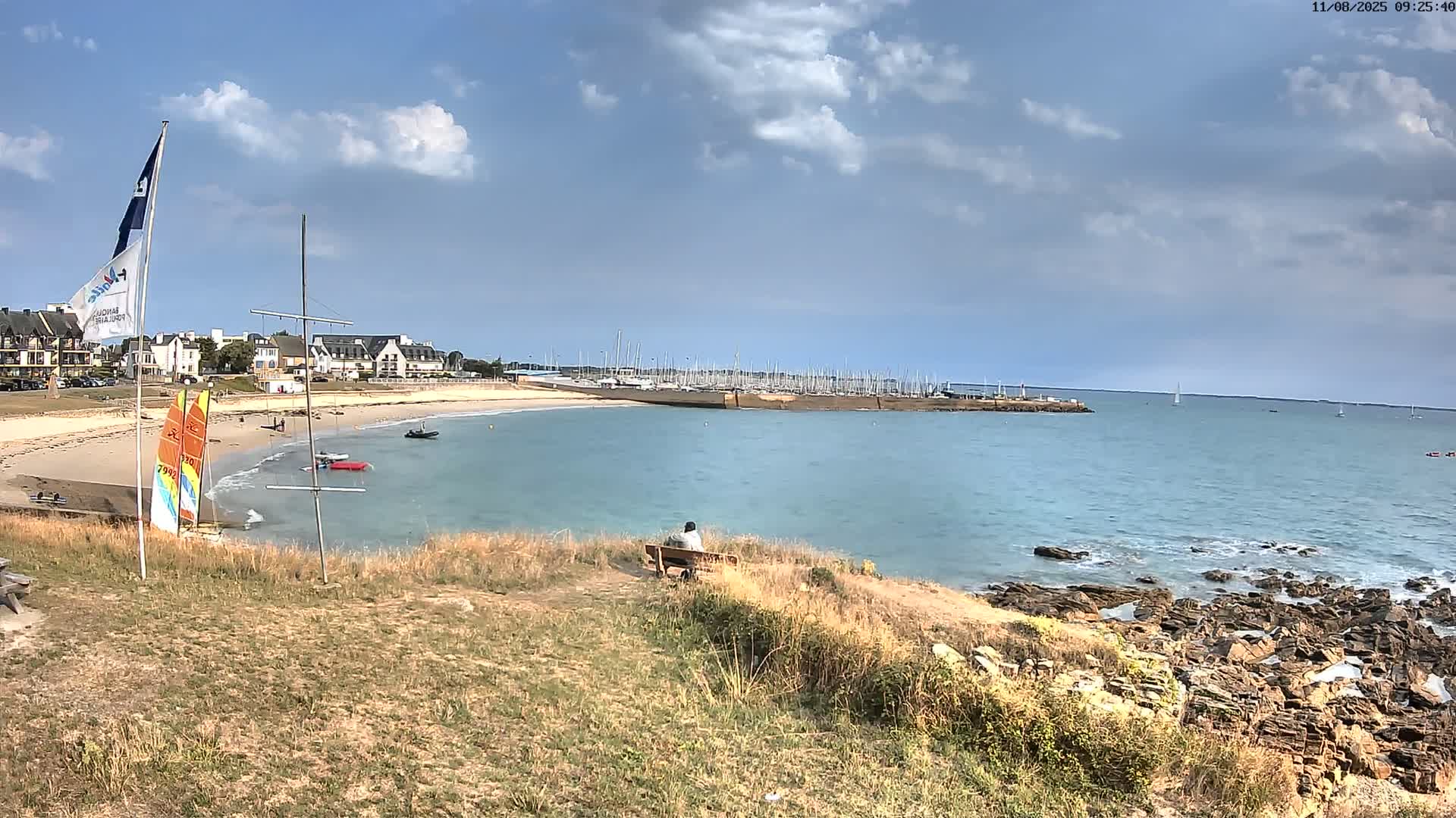 A person sits on a bench overlooking a calm bay with a sandy beach, numerous boats in a marina, and rocky shoreline under a partly cloudy sky.