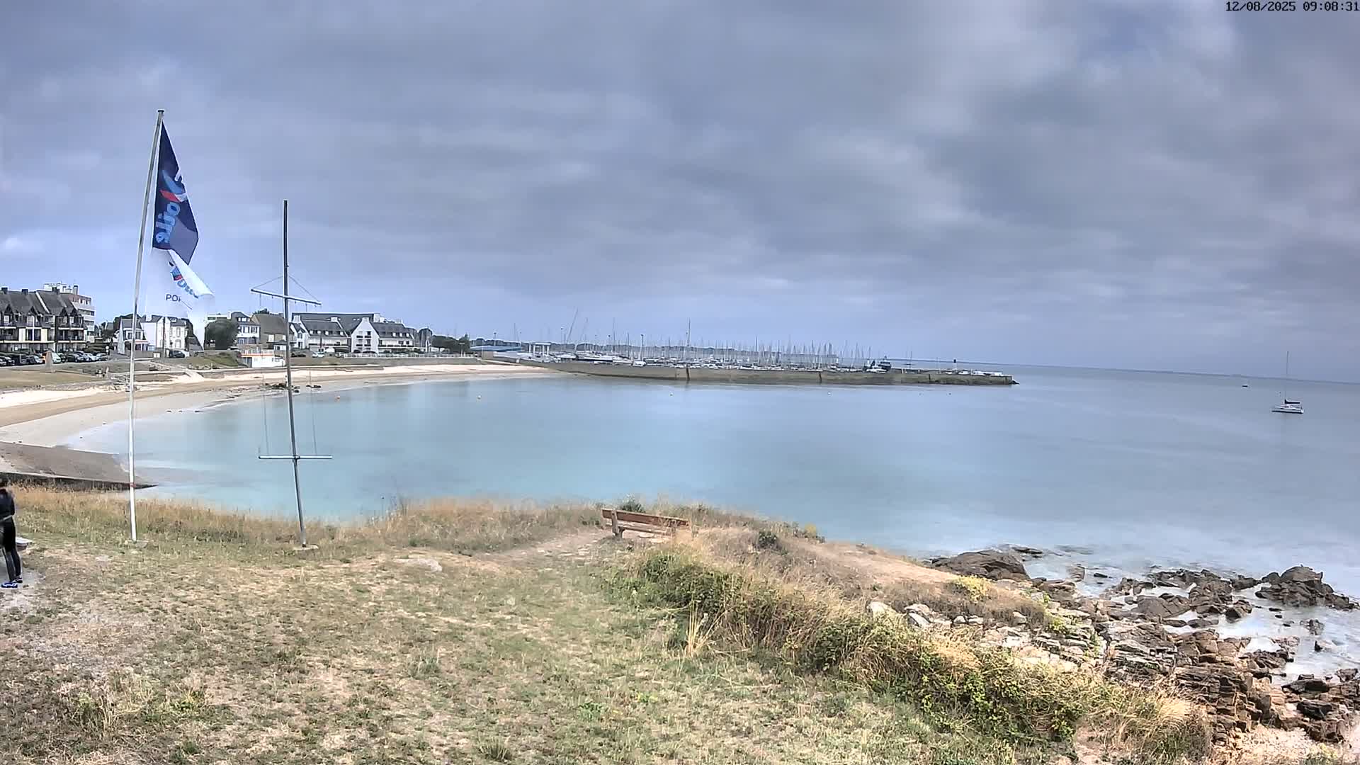 A partly cloudy day reveals a tranquil coastal scene with a sandy beach, calm water, a marina filled with boats, and a rocky shoreline.