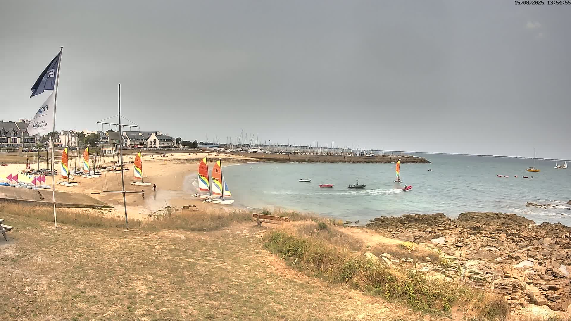 A partly cloudy day reveals a tranquil coastal scene with a sandy beach, calm water, a marina filled with boats, and a rocky shoreline.
