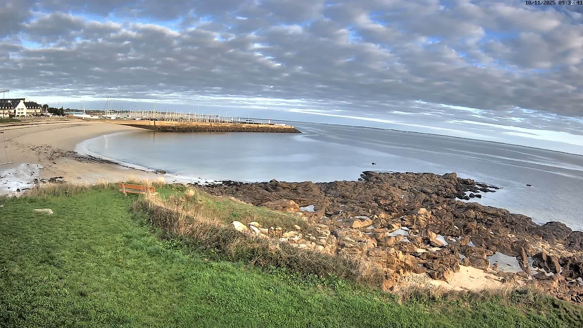 A partly cloudy day reveals a tranquil coastal scene with a sandy beach, calm water, a marina filled with boats, and a rocky shoreline.