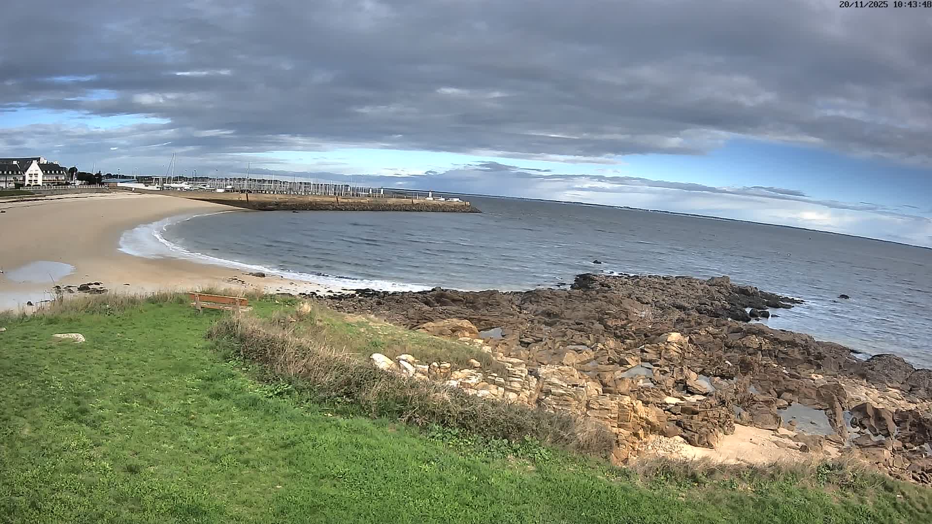 A panoramic coastal scene unfolds under a partly cloudy sky, showcasing a grassy hill and rugged rocky shore in the foreground, leading to a crescent sandy beach with soft waves, a marina filled with sailboats, and distant coastal buildings.