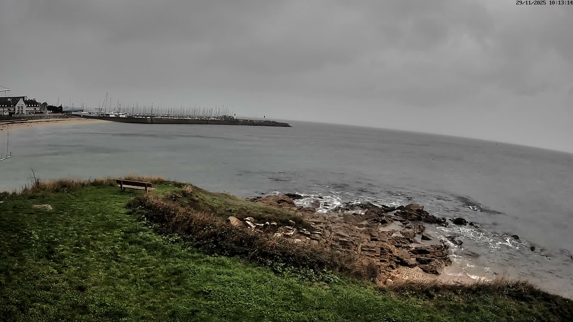 An overcast day overlooks a bay featuring a marina packed with sailboats, a sandy beach, and a rocky coastline with waves crashing against it, all viewed from a grassy clifftop with a lone wooden bench.