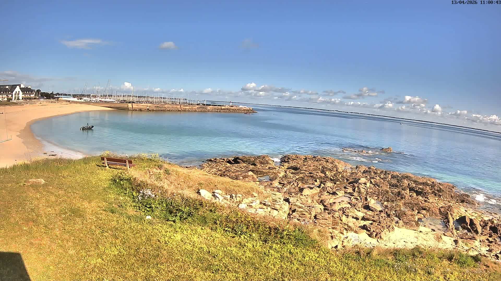 An overcast day overlooks a bay featuring a marina packed with sailboats, a sandy beach, and a rocky coastline with waves crashing against it, all viewed from a grassy clifftop with a lone wooden bench.