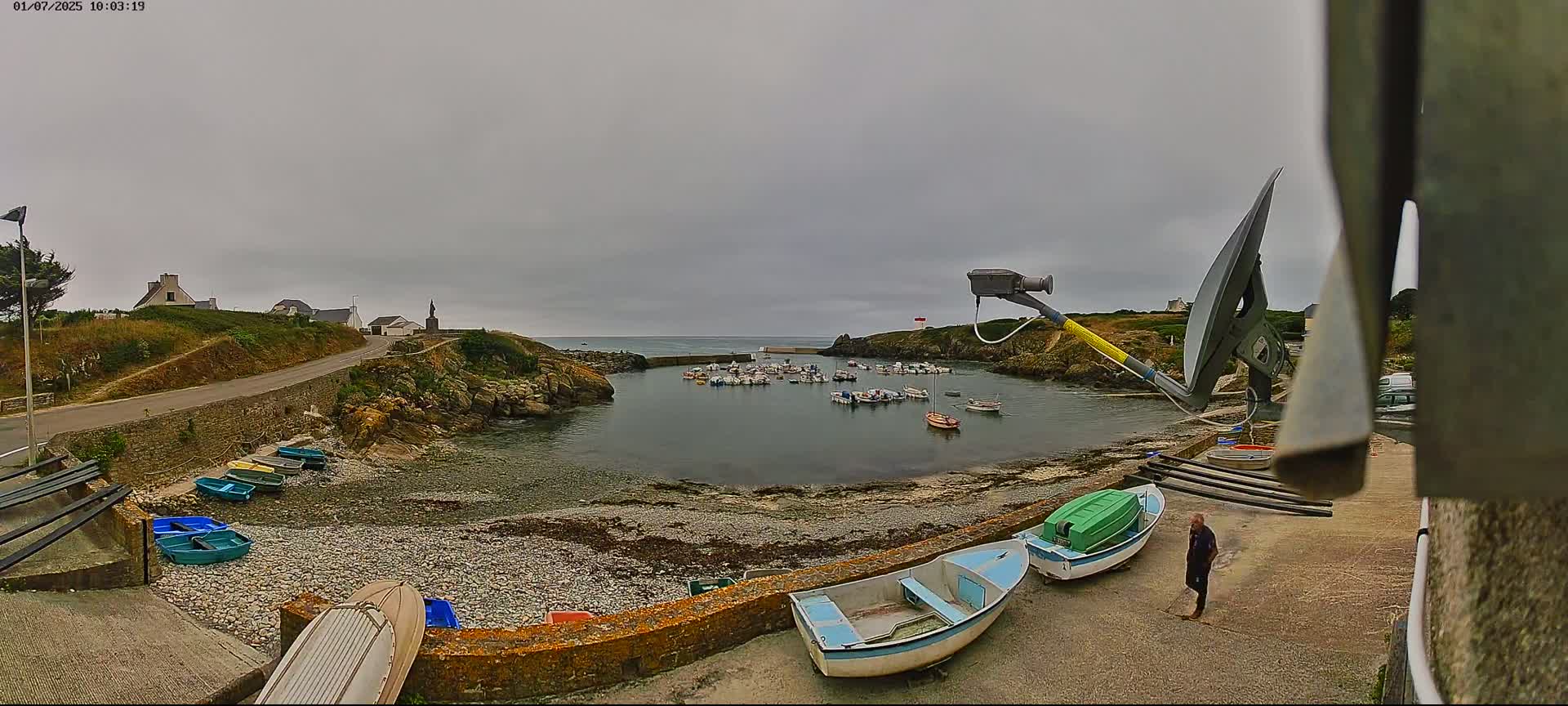 A small harbor filled with numerous boats is nestled between rocky shores under an overcast sky, with a person standing on a nearby paved area.