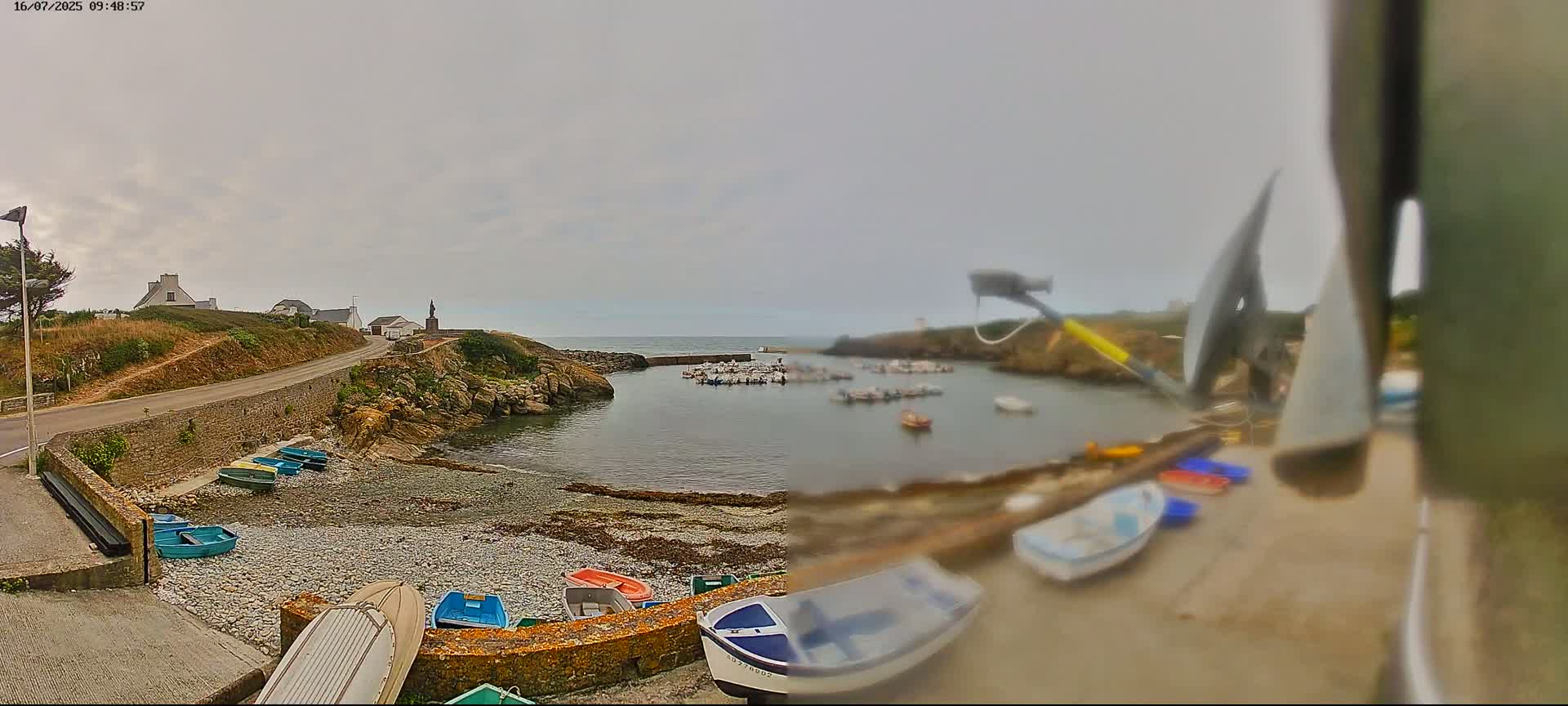 A small, calm harbor with numerous boats is nestled in a rocky cove under an overcast sky, with a road and buildings visible on the adjacent land.