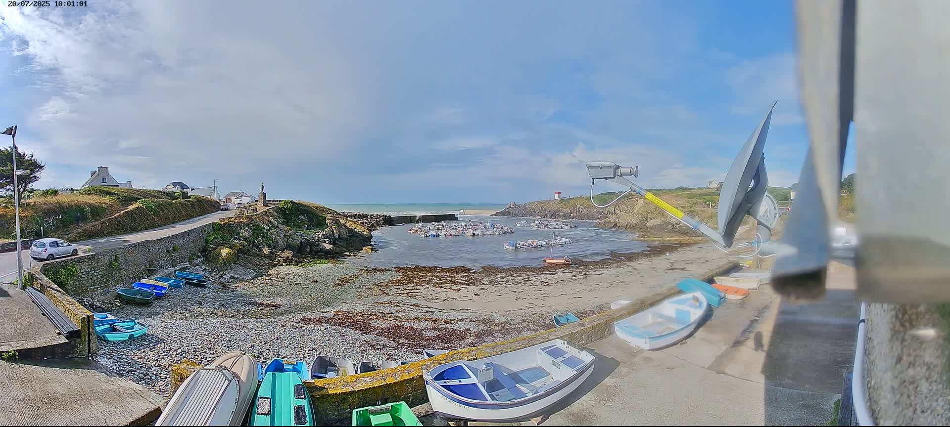 A partly cloudy day reveals a small harbor filled with numerous boats, situated between rocky shores with a road and buildings visible in the background.