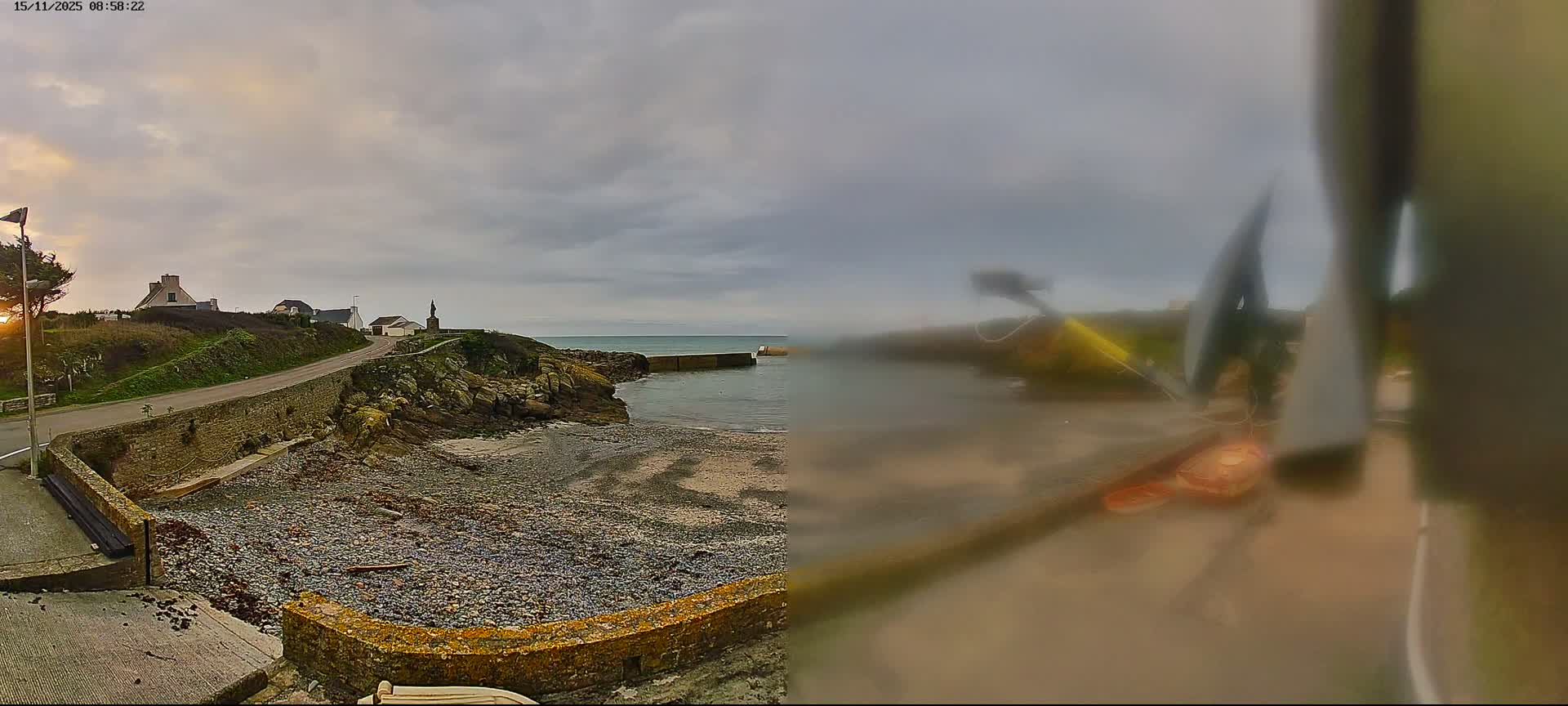 The image displays a split perspective, with a clear view on the left showing a rocky coastal village's winding road, houses, and a pier extending into a calm sea under a partly cloudy dawn sky, while the right side is an extremely blurred and indistinct close-up of abstract shapes.
