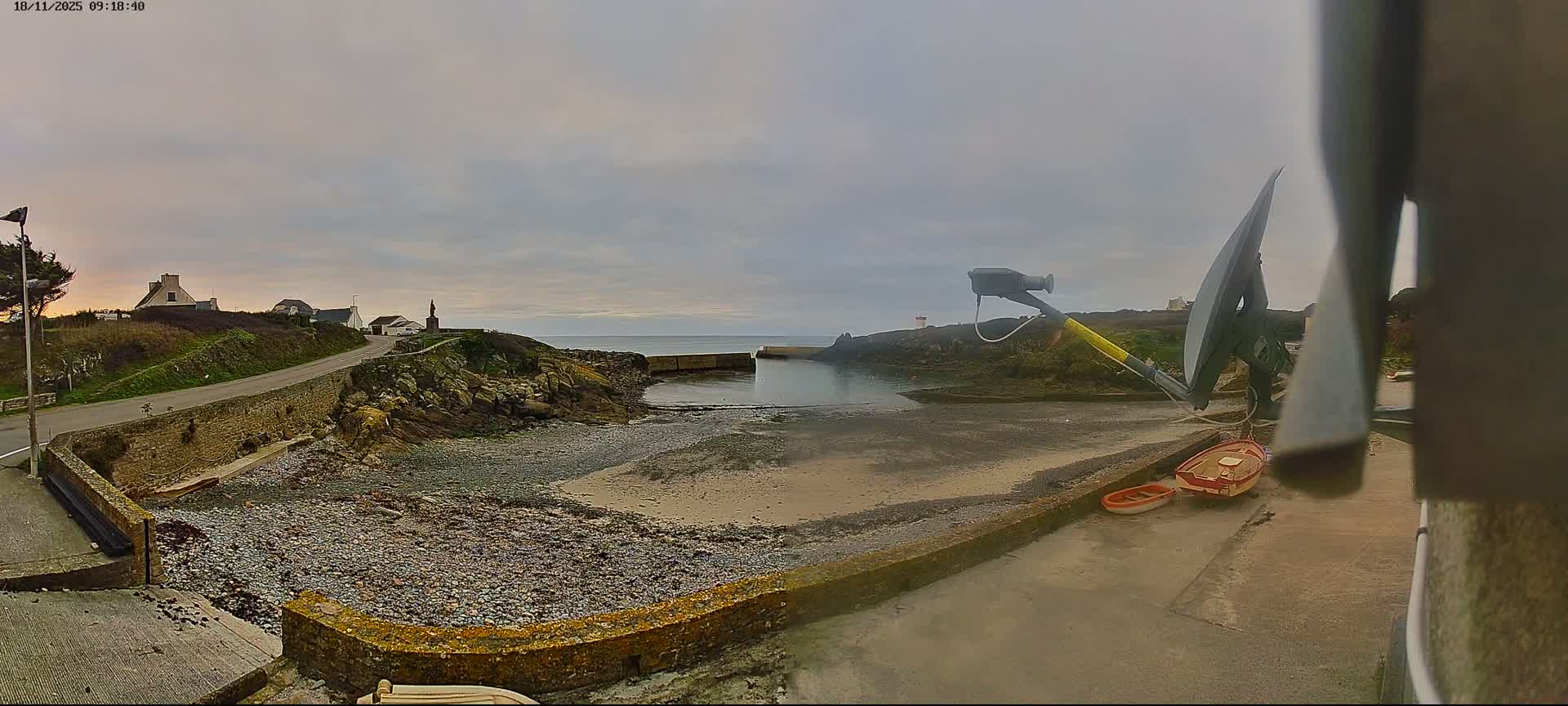 A wide-angle view captures a coastal scene under an overcast sky, featuring a winding road with houses on a hill, a small cove with a rocky beach, and a weather station with two small boats in the foreground.