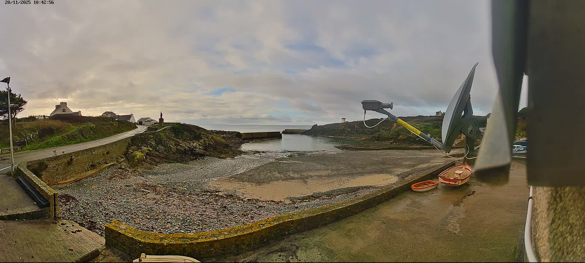 An overcast coastal scene reveals a small, rocky cove at low tide, bordered by a road and houses on a grassy hill, with a large antenna and two small boats visible on a concrete area in the foreground.