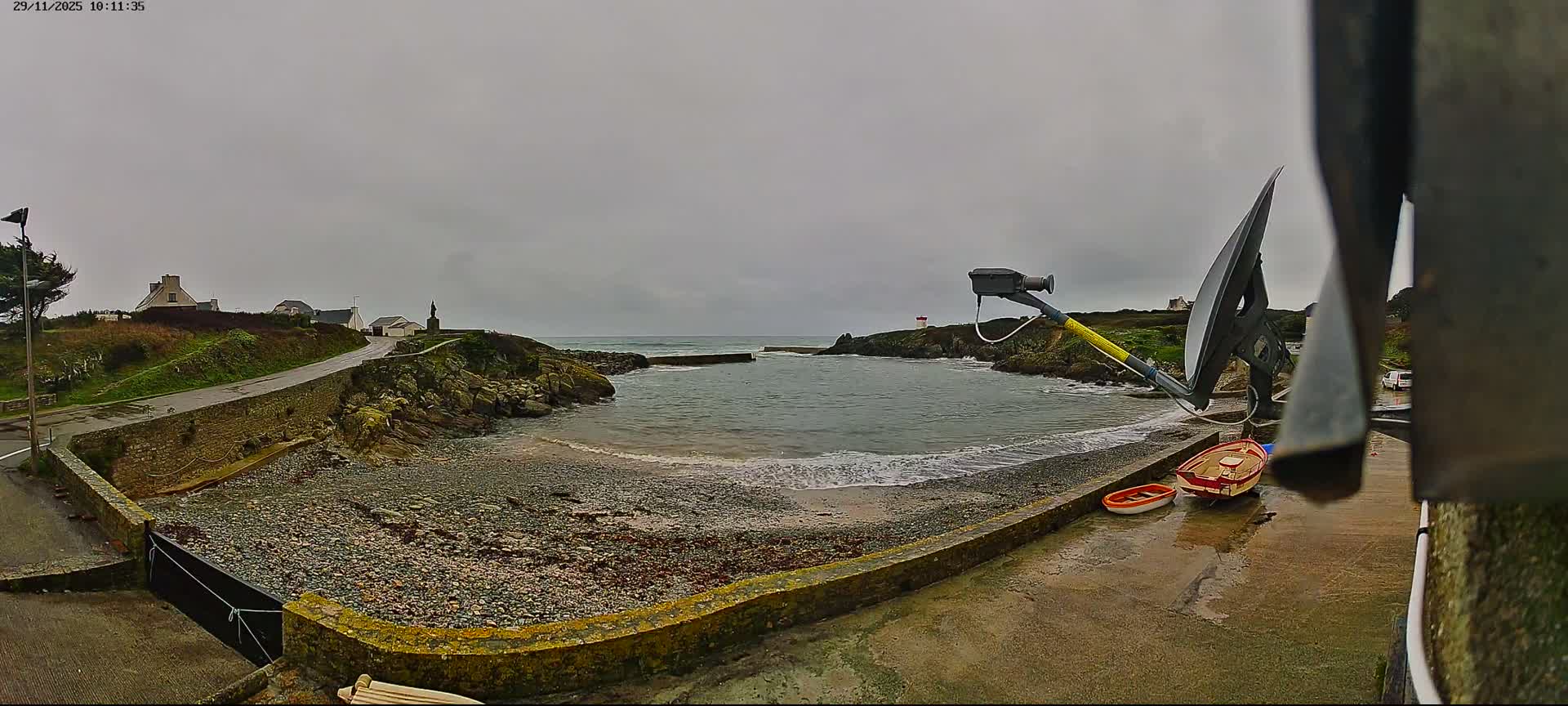 Under a grey and overcast sky, a rocky coastal cove with a small pebbly beach and a jetty is bordered by a winding road and houses on a green hill, with two small boats resting on a wet concrete foreground.
