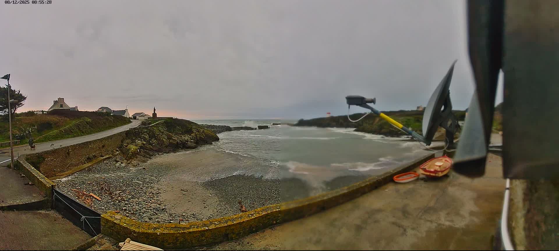 A panoramic view on an overcast day captures a rugged coastline with active waves breaking on a pebble beach and against a distant breakwater, a winding road leading past houses and a person walking a dog, and a prominent statue on a headland.