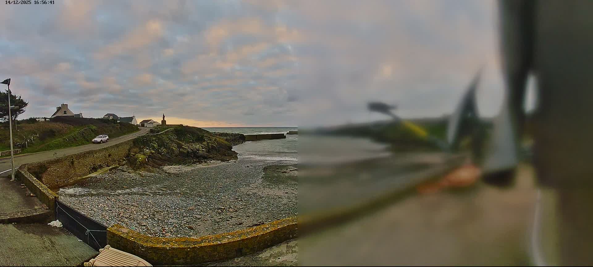 A panoramic view on an overcast day captures a rugged coastline with active waves breaking on a pebble beach and against a distant breakwater, a winding road leading past houses and a person walking a dog, and a prominent statue on a headland.