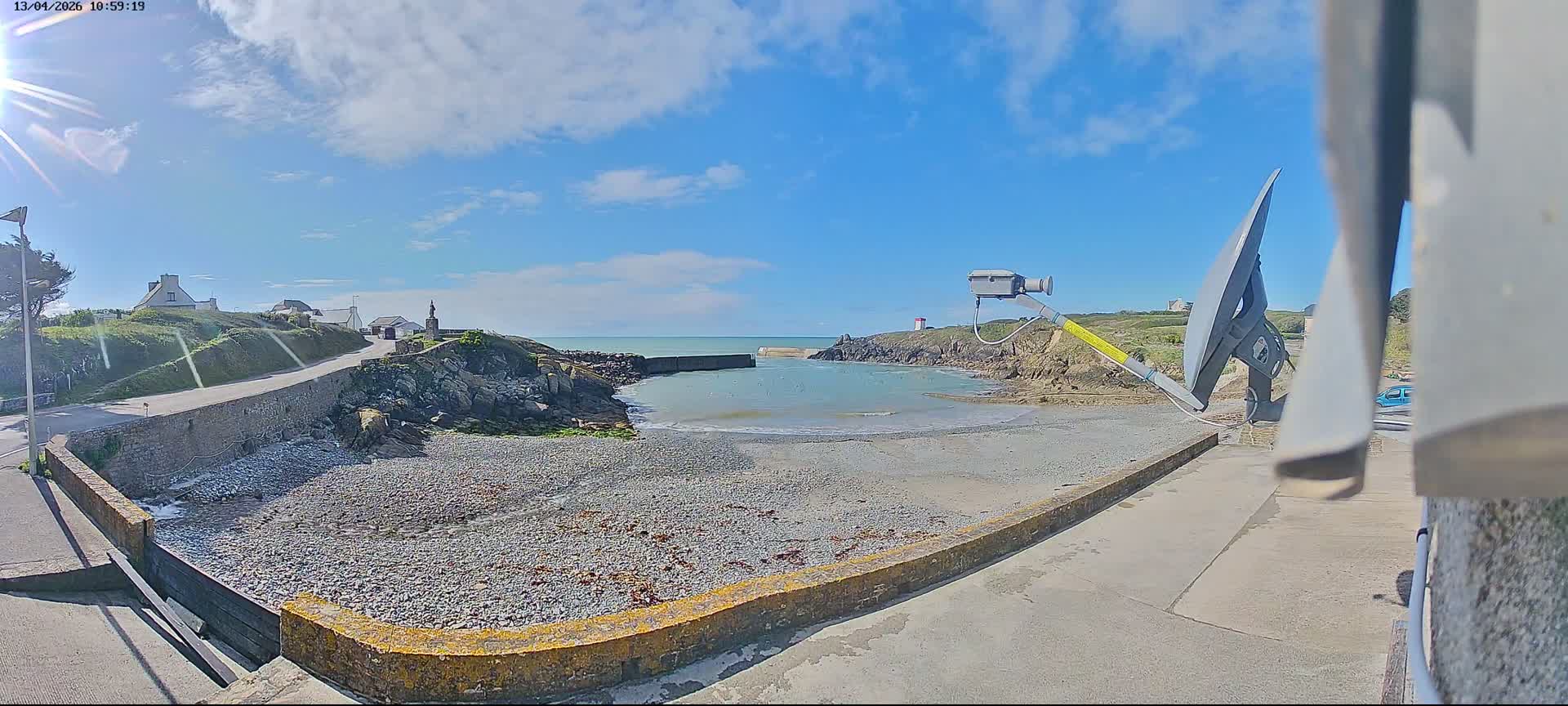A panoramic view on an overcast day captures a rugged coastline with active waves breaking on a pebble beach and against a distant breakwater, a winding road leading past houses and a person walking a dog, and a prominent statue on a headland.