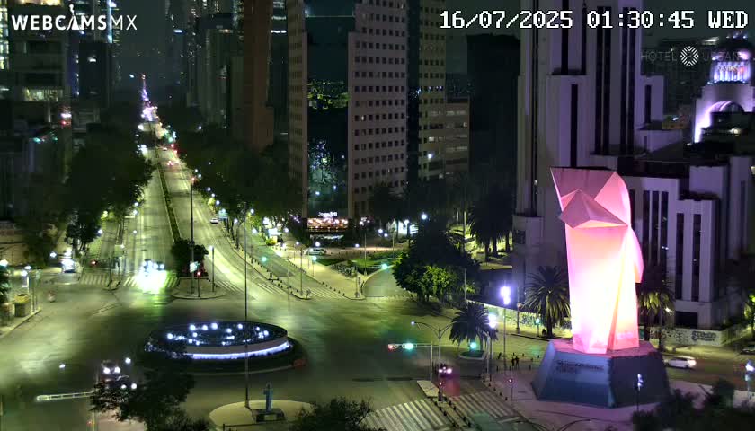 A nighttime aerial view of a city street intersection with sparse traffic, illuminated by streetlights, featuring a large illuminated sculpture in a plaza.