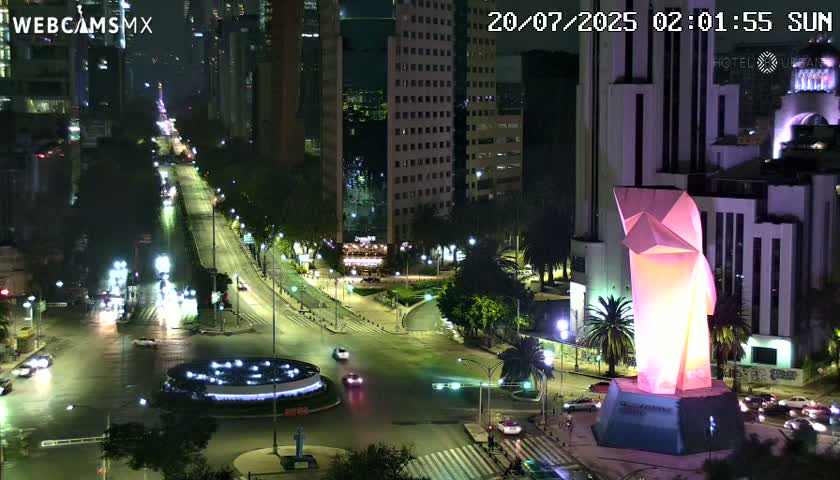 A nighttime, city street scene shows a brightly lit, angular sculpture at a well-lit intersection with vehicles and buildings visible.