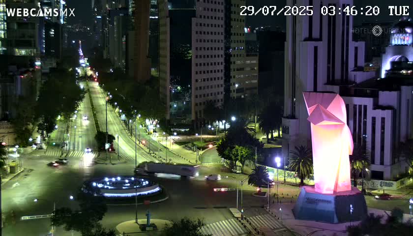 A nighttime, city street scene shows a brightly lit, angular sculpture at a well-lit intersection with vehicles and buildings visible.