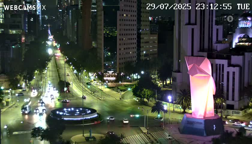 A nighttime, city street scene shows a brightly lit, angular sculpture at a well-lit intersection with vehicles and buildings visible.