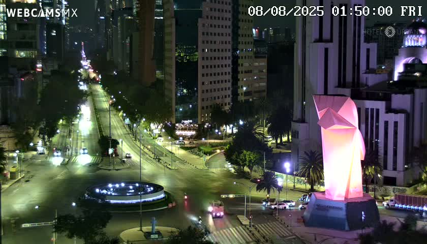 A nighttime, high-angle view shows a city intersection with sparse traffic, illuminated by streetlights, featuring a large, illuminated geometric sculpture near a circular fountain, and tall buildings in the background.