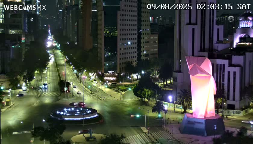 A nighttime, high-angle view shows a city intersection with sparse traffic, illuminated by streetlights, featuring tall buildings and a large, illuminated pink sculpture.