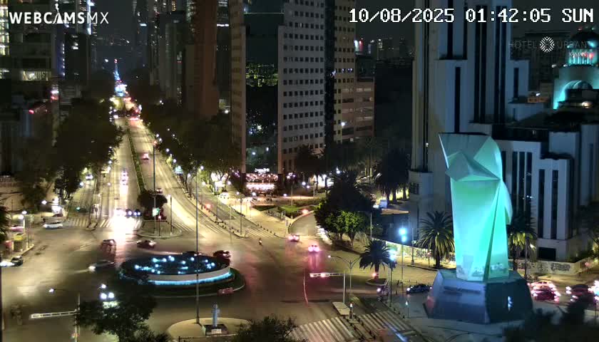 A nighttime, city street scene shows a roundabout with cars driving around it,  tall buildings lining the streets, and a large, illuminated sculpture at an intersection.