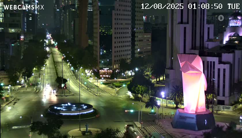 A nighttime, high-angle view shows a city intersection with a large illuminated sculpture, several buildings, and sparsely trafficked roads.