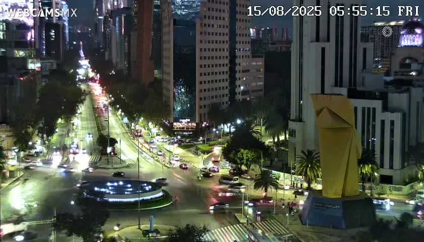 A nighttime, high-angle view shows a city intersection with a large illuminated sculpture, several buildings, and sparsely trafficked roads.