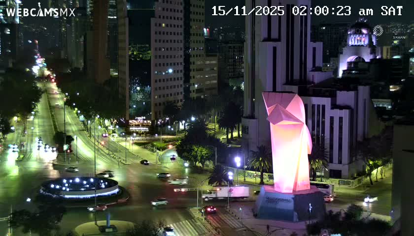 A vibrant city scene at night features a towering, glowing pink and orange modern monument, bustling streets with traffic, a circular illuminated fountain, and numerous brightly lit skyscrapers under a clear sky.