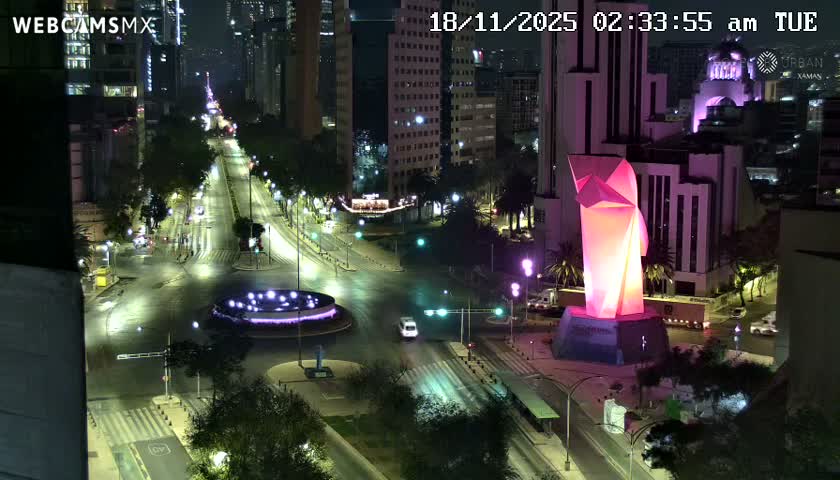 An elevated nighttime view captures a wide city street illuminated by streetlights, featuring a prominent glowing pink geometric sculpture, a circular purple-lit fountain, and distant urban buildings under clear skies.