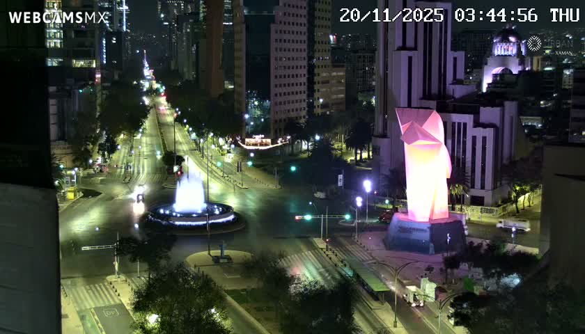 A clear nighttime aerial view captures a bustling city street illuminated by streetlights and vehicle headlights, featuring a bright white fountain and a prominent glowing pink geometric statue, all under clear skies.