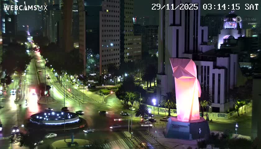 A clear night view captures a vibrant city scene featuring an illuminated pink and orange modern statue, a busy multi-lane boulevard with glowing streetlights and moving cars, and numerous brightly lit urban buildings in the background.