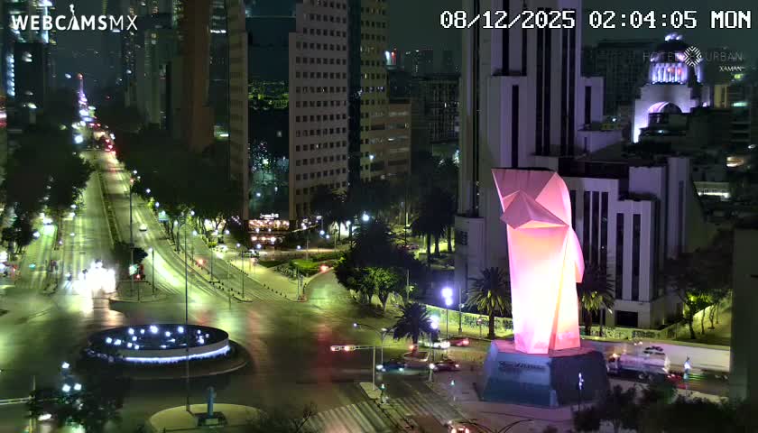 On a clear night, a striking pink-and-white geometric sculpture stands illuminated beside a bustling city street featuring a lighted fountain, numerous cars, and tall buildings in the background.