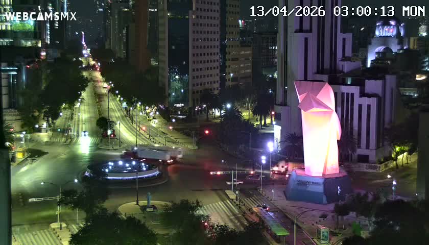 On a clear night, a striking pink-and-white geometric sculpture stands illuminated beside a bustling city street featuring a lighted fountain, numerous cars, and tall buildings in the background.