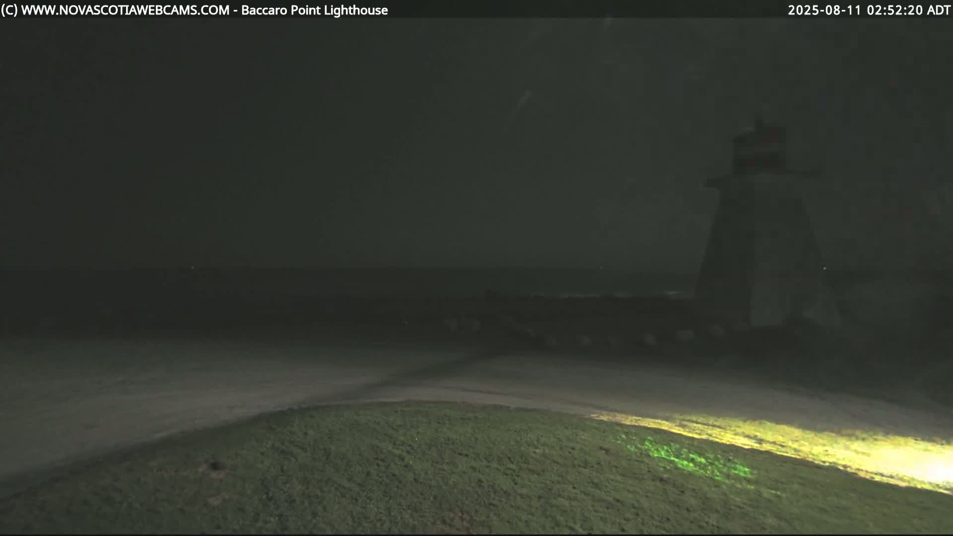 A dark, nighttime scene shows a lighthouse standing on a grassy hill overlooking a dark body of water under a somewhat hazy sky.
