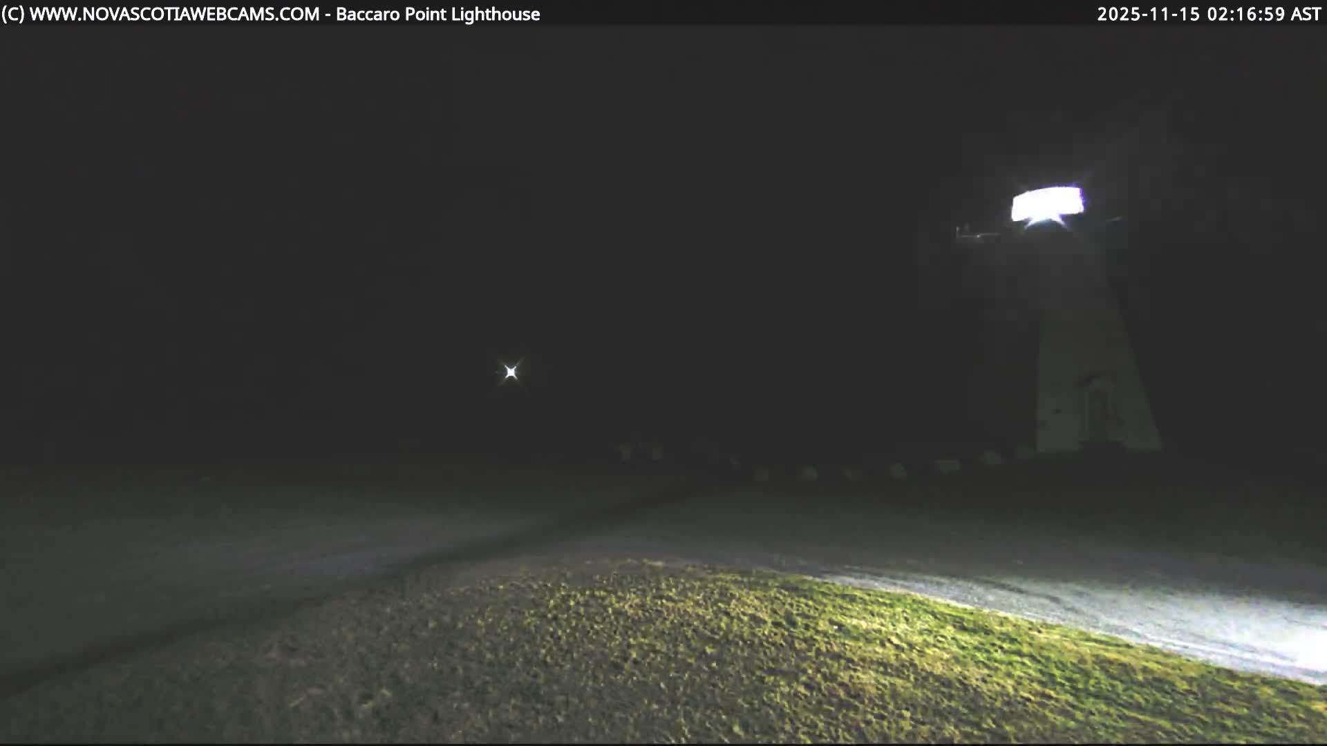 A clear, dark night scene reveals a brightly lit lighthouse on the right, a distant star-like light on the left, and a road flanked by illuminated grassy areas in the foreground.