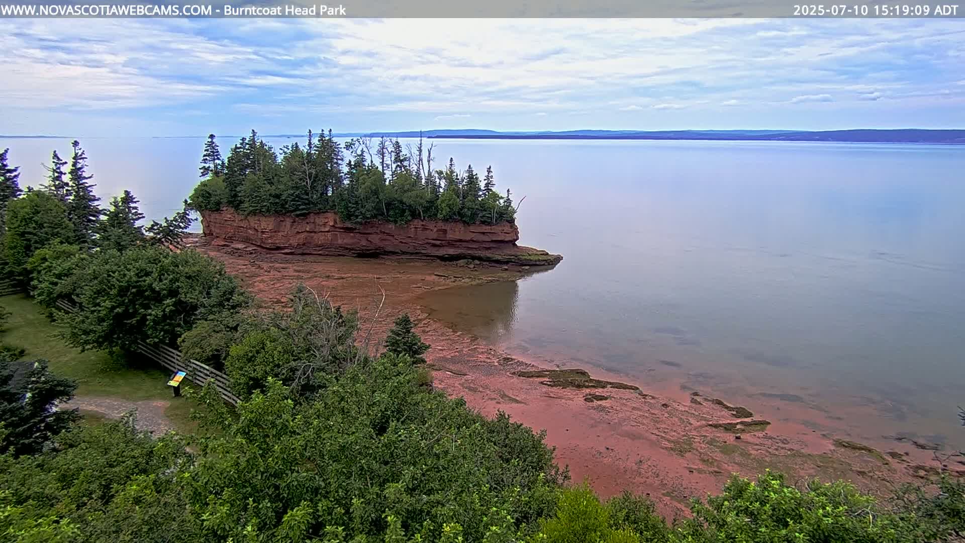 A red-rock island sits in calm, light-grey water under a partly cloudy sky, surrounded by green trees and a red sandy shoreline.