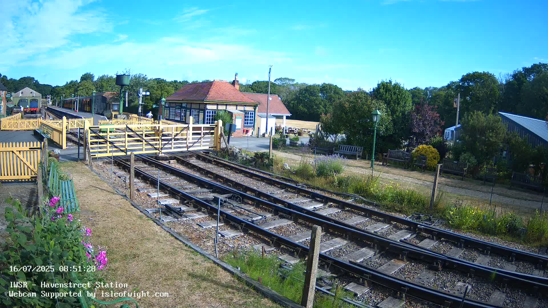 A sunny day at a quaint train station with multiple tracks, a small station building, and benches along a grassy area.