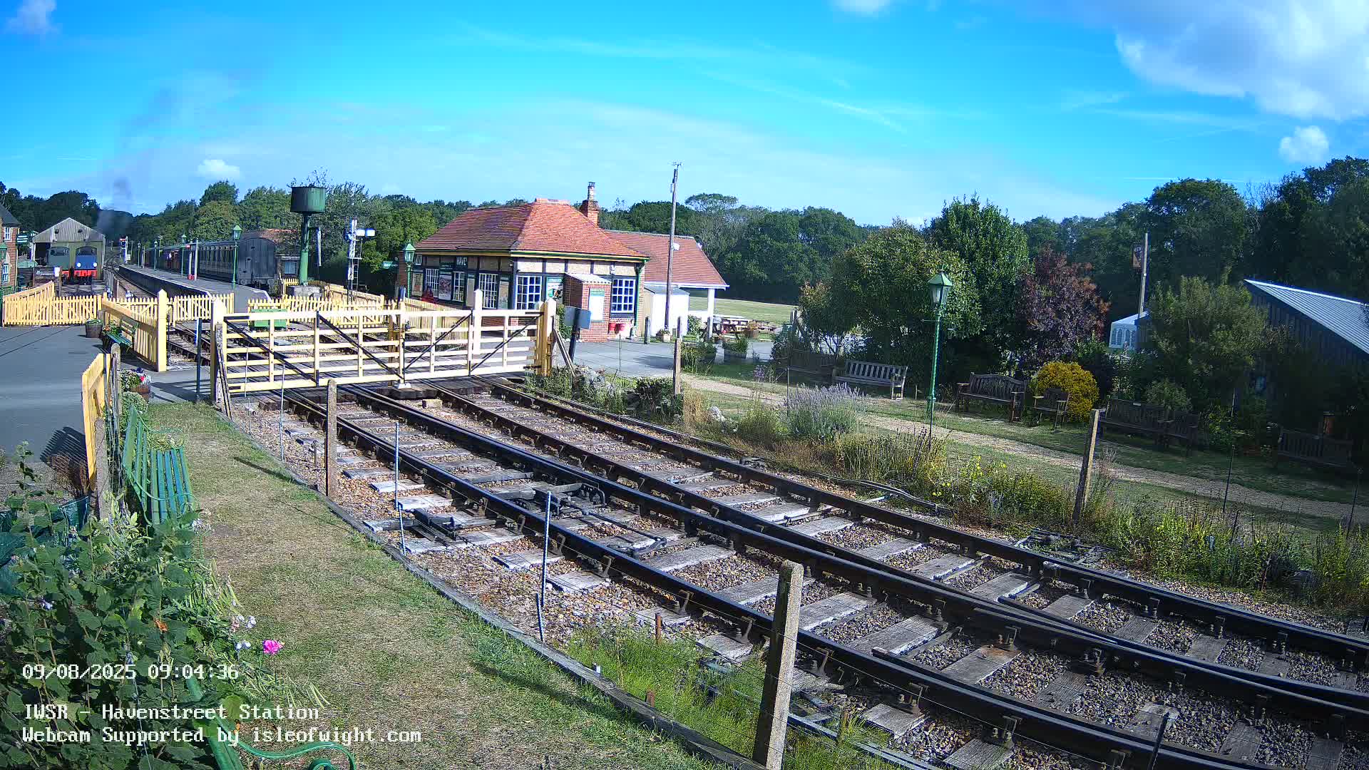 A train station with a small building and railway tracks is shown under sunny skies with some clouds.